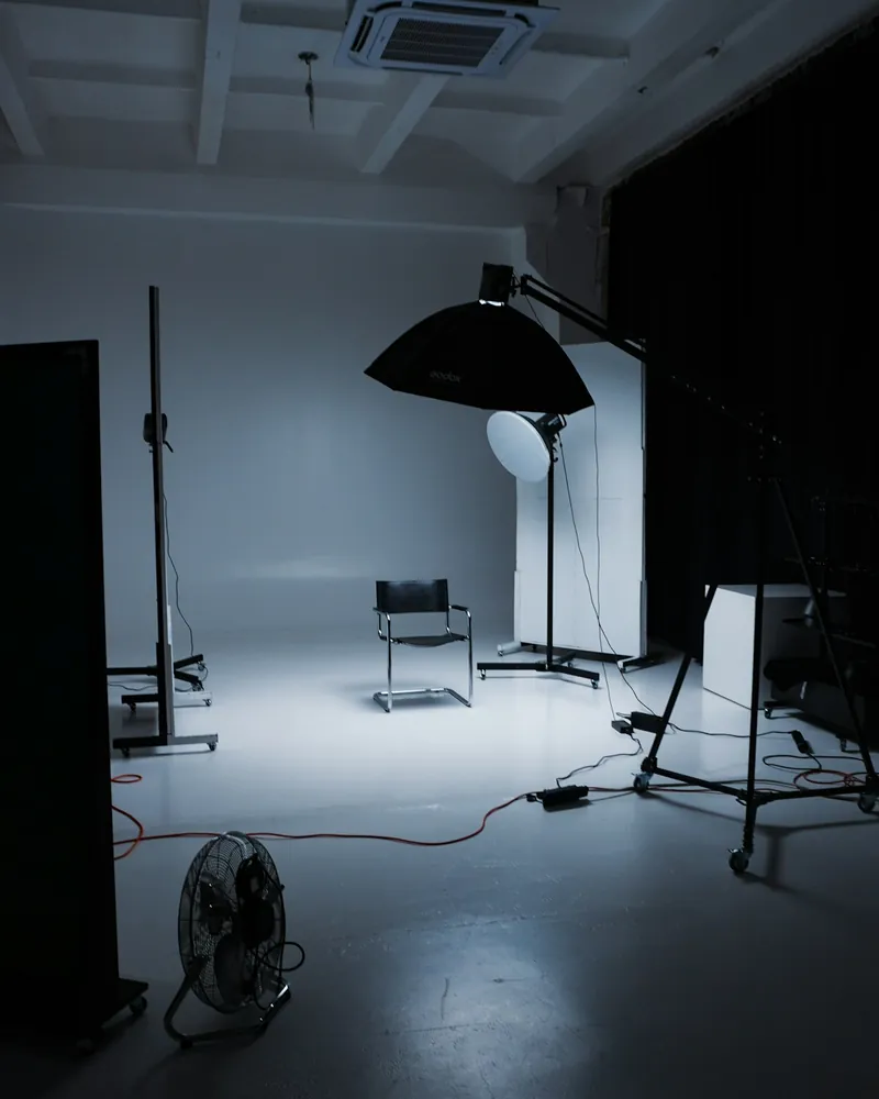Estúdio em penumbra com uma cadeira vazia sobre um fundo branco contínuo, uma softbox suspensa num braço e um beauty dish num tripé